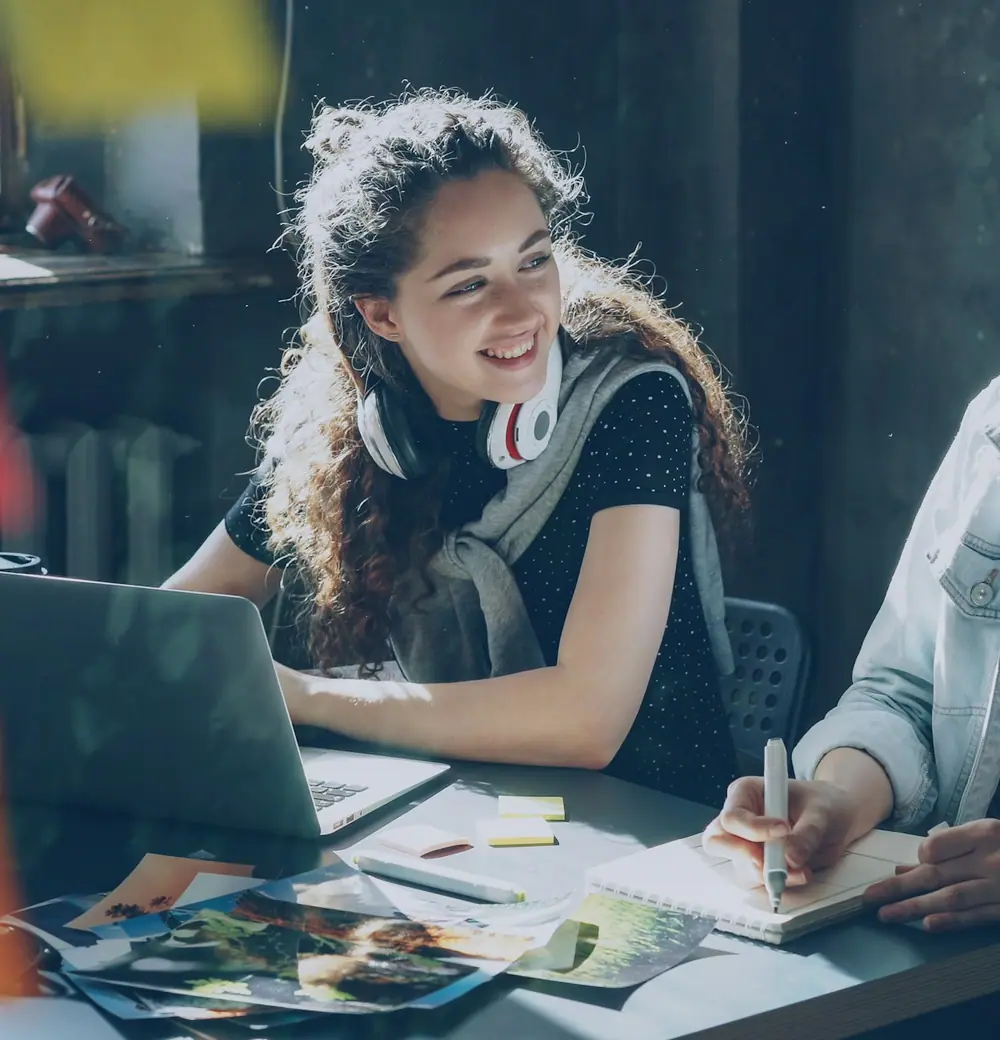 Two women at a desk with a laptop and notes, smiling as they work