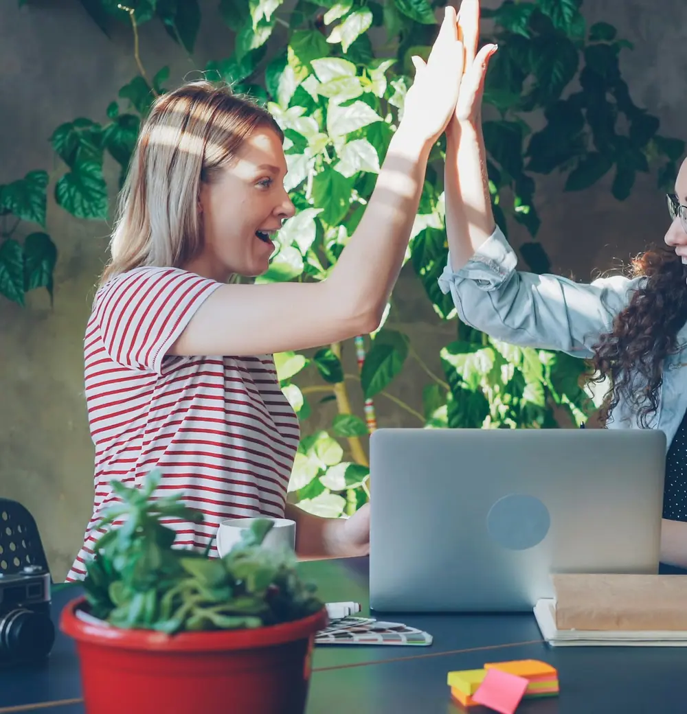 Two women high-fiving across a desk with a laptop open