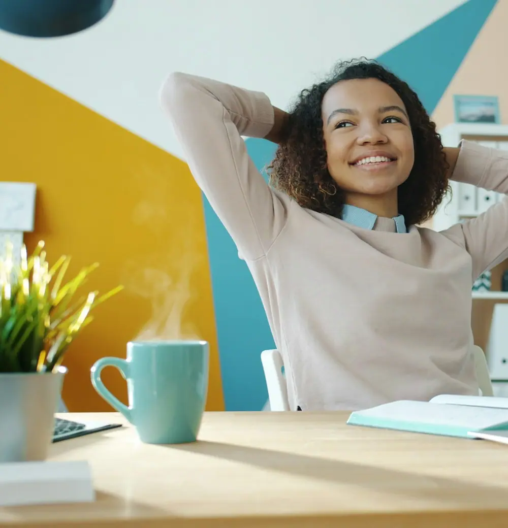 A woman leaning back in her chair, hands behind her head, smiling at her laptop