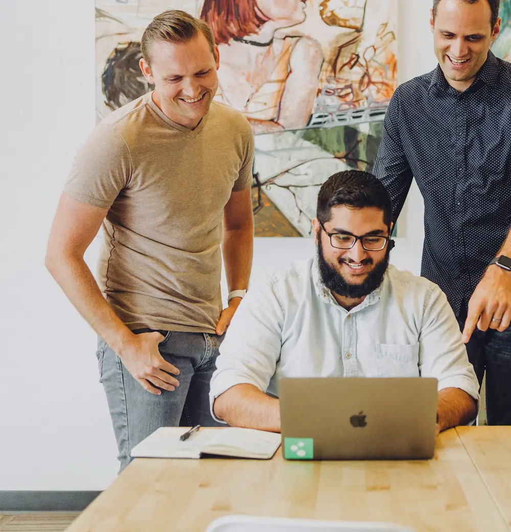 A small team gathered at a laptop, one man pointing at the screen while the others smile