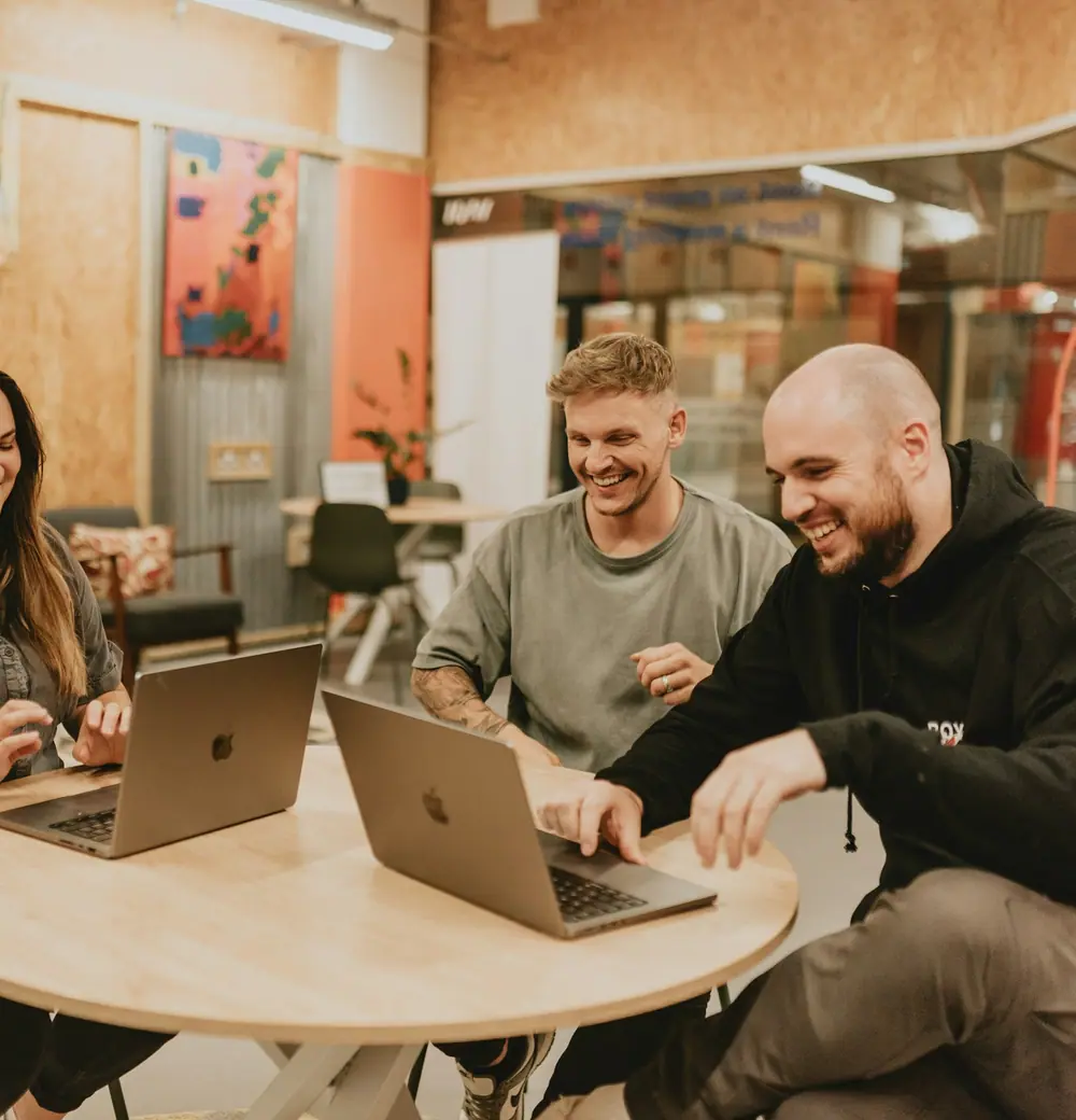 Three people laughing together around laptops at a round table
